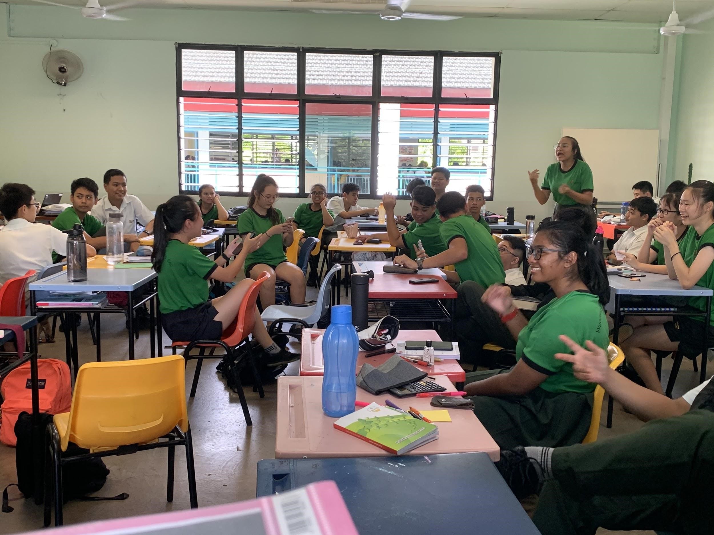 Students in green uniforms sit at desks in a classroom while a teacher stands and speaks at the back.